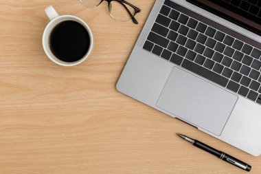 Üst Manzara Ahşap ofis masası. Göz gözlüğü, kahve fincanı, dizüstü bilgisayar, ahşap masa arkasında kalem ofis malzemeleri olan düz bir çalışma alanı.