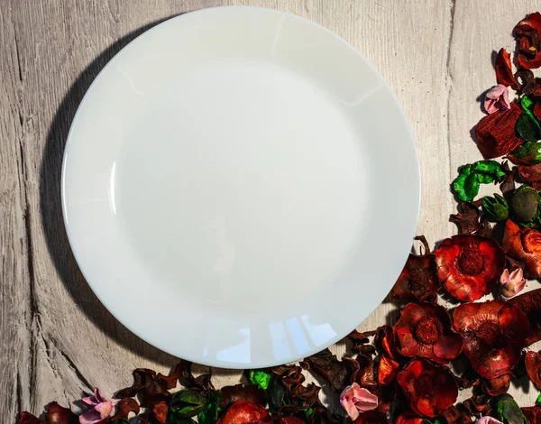 Ahşap arka plan boş beyaz tabakta kuru çiçek yaprakları ile süslenmiş. Yukarıdan görüntülemek.