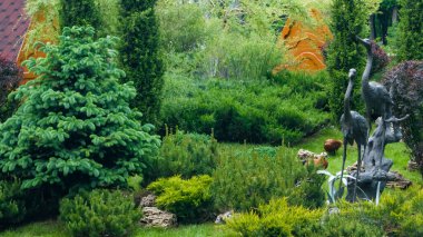 Parktaki yeşil çimenlerin ve ağaçların arasında kahverengi ördekler. Leylek metal heykelleri. Arkaplan