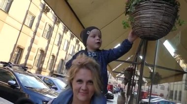 Prag, Çek Cumhuriyeti - 7 Mayıs 2018: Çocuk annesinin boynuna oturuyor. Akşam güneş ışınlarının sokakta yürüyorlar. Ağır çekim