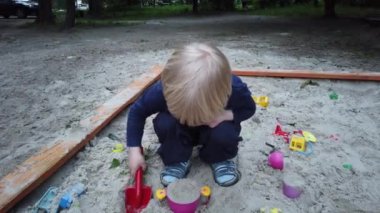 Üç yaşında bir çocuk kum havuzunda oynuyor. Rebony bahçede kumla oynuyor. Kum havuzunda çocuk oyuncakları var. Çocuk spatula ve kova.