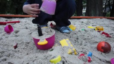 Üç yaşında bir çocuk kum havuzunda oynuyor. Rebony bahçede kumla oynuyor. Kum havuzunda çocuk oyuncakları var. Çocuk spatula ve kova.