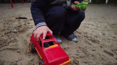 Üç yaşında bir çocuk kum havuzunda oynuyor. Rebony bahçede kumla oynuyor. Kum havuzunda çocuk oyuncakları var. Çocuk spatula ve kova.