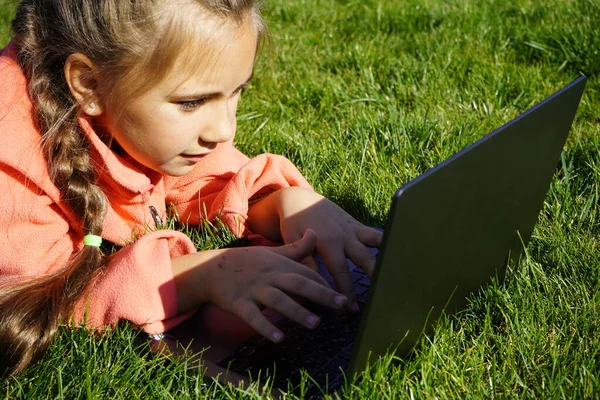 Ein siebenjähriges Mädchen arbeitet auf einem Laptop im grünen Gras. — Stockfoto
