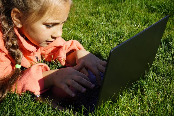 Ein siebenjähriges Mädchen arbeitet auf einem Laptop im grünen Gras. — Stockfoto