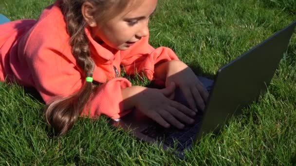 一个7岁的女孩在绿草上的笔记本电脑上工作 远程学习 在松树背景上拿着笔记本电脑的女孩 街上有一台电脑的女孩 草地上的孩子玩电脑游戏 远程教育 — 图库视频影像