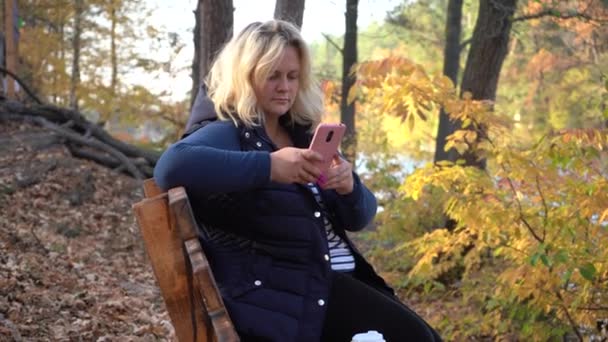 ベンチに座っている女の子は 湖と秋の森の背景にスマートフォンを使用しています 湖の前の秋の森のベンチに女の子が座っている — ストック動画