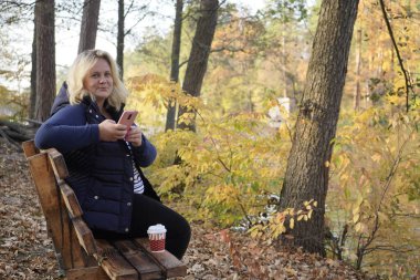 Bankta oturan kız arka planda akıllı telefon kullanıyor.