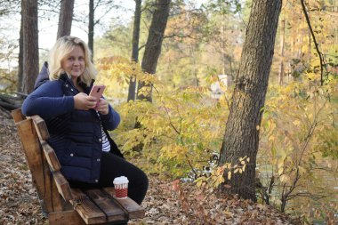 Bankta oturan kız arka planda akıllı telefon kullanıyor.