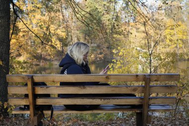 Bankta oturan kız arka planda akıllı telefon kullanıyor.