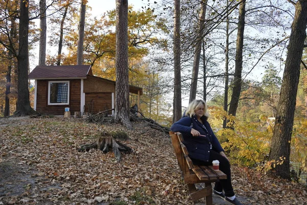 Uma menina se senta em um banco em meio a uma cabana na floresta de outono e bebe — Fotografia de Stock