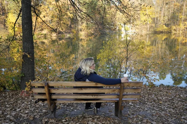 A girl sits on a bench against the background of a lake and autu — Stock Photo, Image