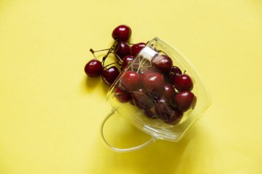 Şeffaf bardaktaki kirazlar sarı arka planda yatıyor.