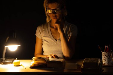 Bir kız karanlık bir odada lambanın ışığında oturuyor ve ödev, ödev, ödev yapıyor.