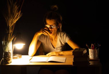 Bir kız karanlık bir odada lambanın ışığında oturuyor ve ödev, ödev, ödev yapıyor.