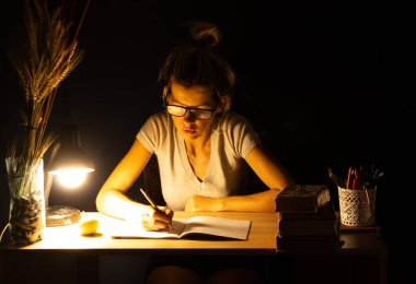 Bir kız karanlık bir odada lambanın ışığında oturuyor ve ödev, ödev, ödev yapıyor.