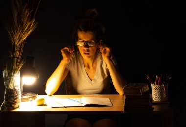 Bir kız karanlık bir odada lambanın ışığında oturuyor ve ödev, ödev, ödev yapıyor.