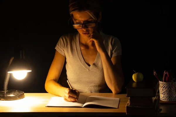 a girl sits at a table by the light of a lamp in a dark room and does homework, homework, homework