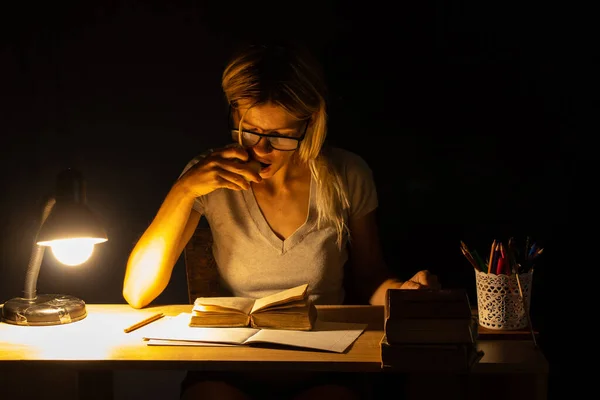 a girl sits at a table by the light of a lamp in a dark room and does homework, homework, homework