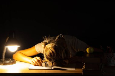 Karanlık bir odada, lambanın ışığında uyuyan, öğrenen, aşırı zorlayan bir kız.