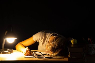 Karanlık bir odada, lambanın ışığında uyuyan, öğrenen, aşırı zorlayan bir kız.