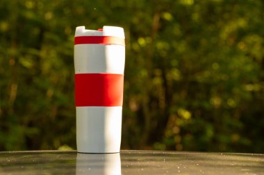 Beyaz termo bardak, ormanda güneşin altında bulanık bir arkaplan üzerinde duruyor.