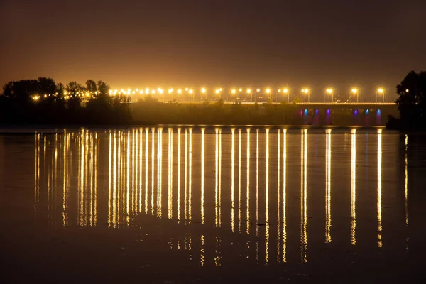 夜晚，在灯柱的灯光下，长距离的暴露在水中，映照在水面上的一座桥 — 图库照片