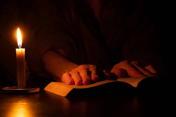grandmother\'s hands lie on an open book on the table by the light of a candle in the dark, reading a prayer with a candle