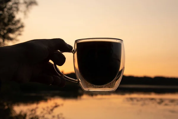 Ręka Trzyma Przezroczystą Szklaną Filiżankę Kawy Zachodzie Słońca Tle Rzeki — Zdjęcie stockowe