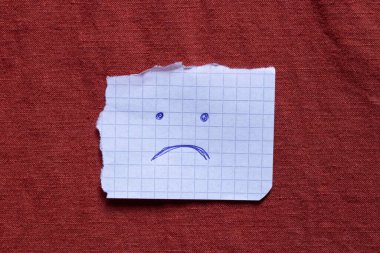 Mavi kalemle çizilmiş üzgün bir yüzü olan kare bir kağıt parçası, duygu, ruh hali ve üzüntünün ifadesi olarak kırmızı bir kumaşın üzerinde yatar.