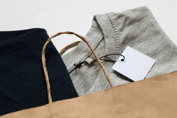 Gri ve mavi etiketli iki tişört yakın plan tasarım için beyaz arka planda bir kese kağıdında duruyor.
