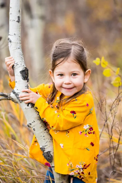 Little girl in yellow shirt posing from behind a small tree