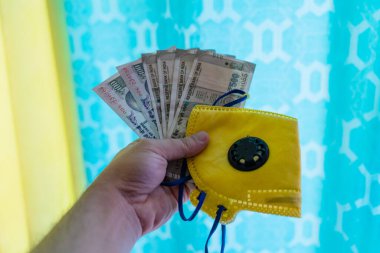 El ele tutuşan Hint banknotları, sarı maske ve dezenfektan, renkli bir arka plana karşı