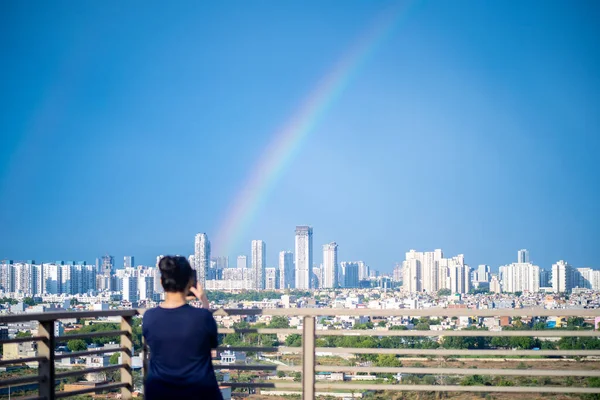 Young indian girl wearing hair in a bun and with blue dress photographing rainbow over gurgaon delhi noida cityscape on a monsoon day
