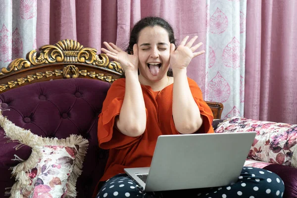 Young indian woman in a red dress sitting cross legged on an ornate sofa with elegant curtains working on a laptop working from home in the coronavirus pandemic lockdown