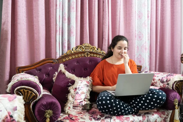 Young indian woman in a red dress sitting cross legged on an ornate sofa with elegant curtains working on a laptop working from home in the coronavirus pandemic lockdown