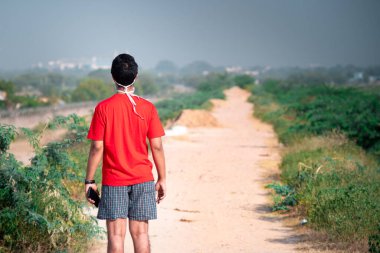 Kırmızı tişörtlü ve şortlu genç Hintli çocuk uzun bir toprak yola bakıyor. Kariyerinde ve çalışmalarında kendini adamış ve mesafe katediyor.