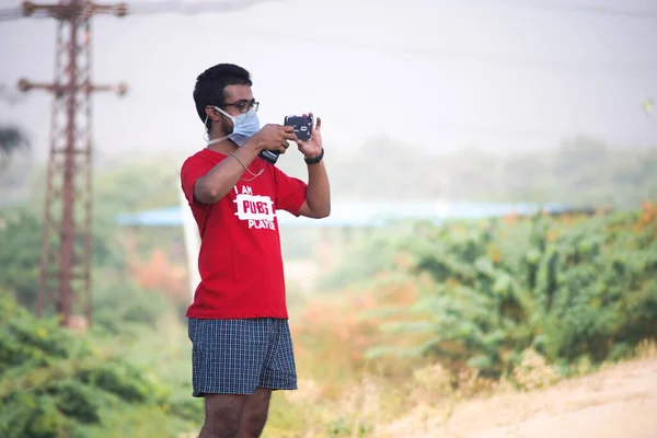Tânăr indian băiat în cămașă roșie PUBG și pantaloni scurți folosind un telefon mobil pentru a face o fotografie cu fundal rural neclar cu copaci și un turn de electercity în spatele lui — Fotografie, imagine de stoc