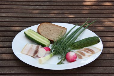 Tuzlu pastırma, çavdar ekmeği ve taze sebzeler, yeşil soğan, kırmızı turp, yeşil dereotu, yeşil bir bahçeye karşı ahşap bir masa üzerinde bir tabak salatalık