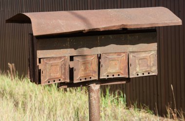 Kahverengi metal bir çitin arkasında paslı posta kutuları var.