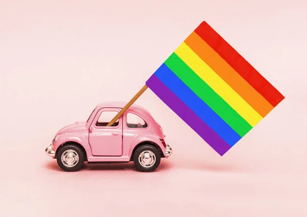 Pink retro toy car delivering bright rainbow gay flag on soft pink backgraund. Concept of gay parade, LGBT community and celebrating Pride