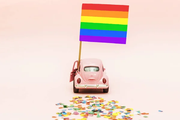 Pink retro toy car delivering bright rainbow gay flag on soft pink backgraund leaving a trail of colored confetti. Concept of gay parade, LGBT community and celebrating Pride