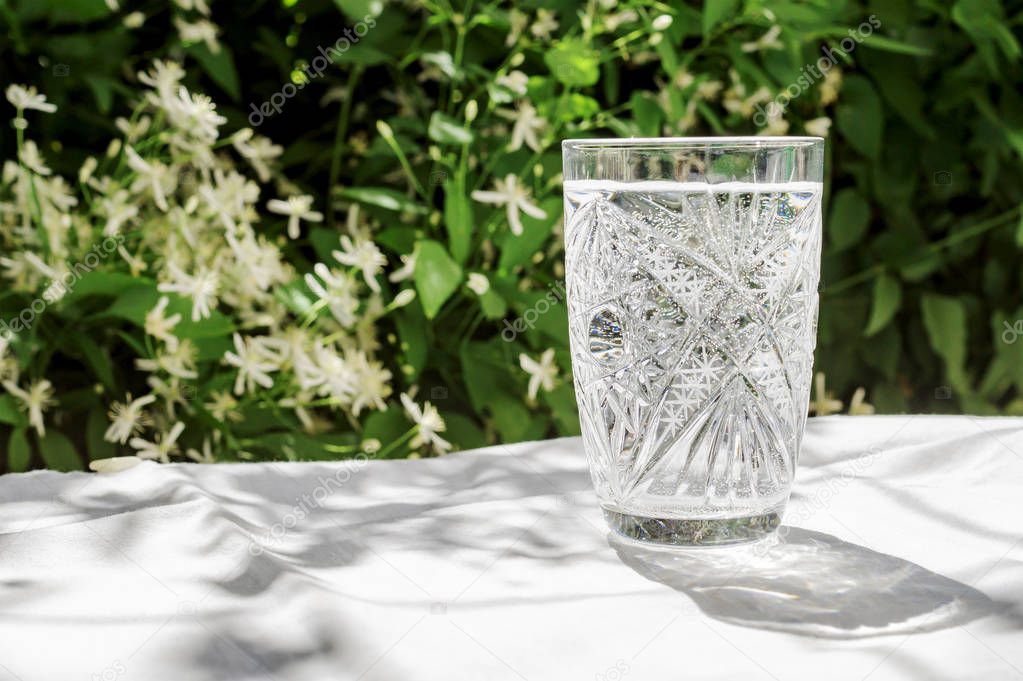 A glass of clean water with bubbles stands on a table with a white cloth against the background of blurred flowers. Copy space for your text. The concept of water and a healthy lifestyle