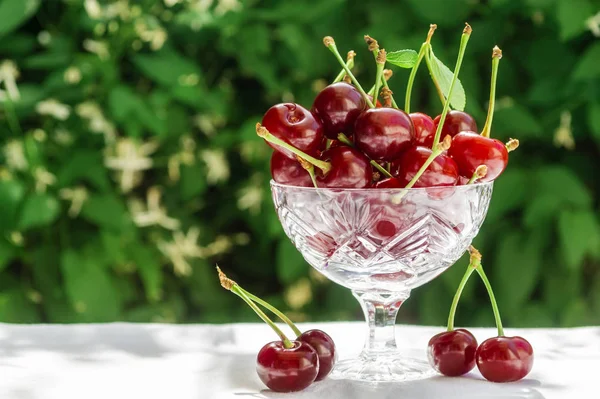 Cerises fraîches dans un bol en cristal sur une table avec un chiffon blanc sur un fond naturel vert de fleurs floues. Concept d'aliments sains, vitamine C. Copiez l'espace pour votre texte — Photo