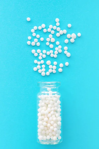 Homeopathic globules in glass bottle on pastel blue background. The globules are scattered from bottle. Alternative homeopathy medicine herbs, healthcare and pills concept. Selective focus, vertical