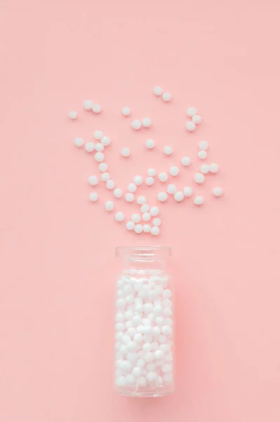 Homeopathic globules in glass bottle on pastel pink background. The globules are scattered from bottle. Alternative homeopathy medicine herbs, healthcare and pills concept. Selective focus