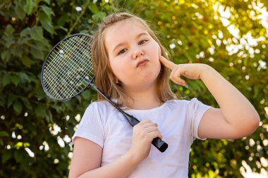 Küçük bir kız şakacı bir şekilde büyüleyici bir yüz ifadesi takınıyor. Sıcak ve güneşli bir yaz gününde dışarıda badminton oynayan tatlı küçük bir kız. Çocuklarla açık hava aktiviteleri. Yaz eğleniyor. Boşluğu kopyala.