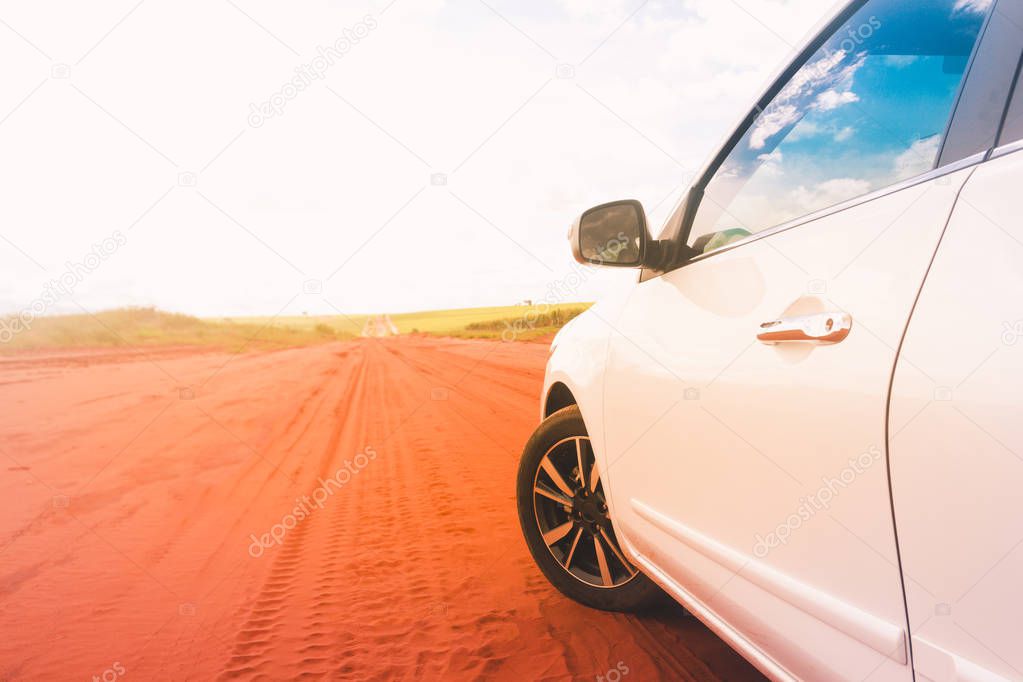 Picture of the front side of a white sports car, the wheel appearing and with reflections of the sun on the driver's glass. Dirt road. Space for text.