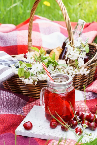 Zumo de cereza en tarro de albañil y cesta de picnic con comida, flores . — Foto de Stock