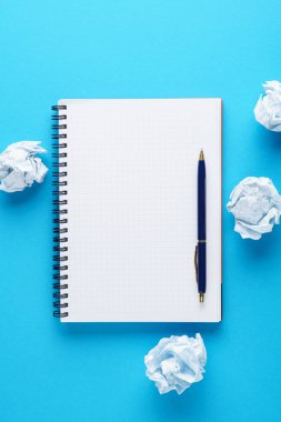 Mavi arka plan üzerinde açık defter, kalem ve buruşuk kağıt topları.